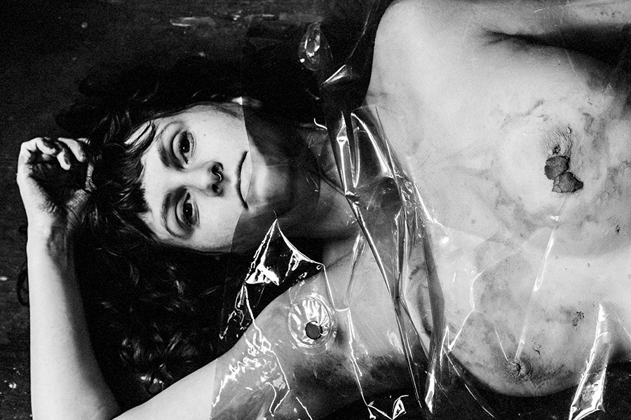 Liegende Frau unter transparenter Folie, Blick direkt in die Kamera, Ton auf Haut und Folie sichtbar.