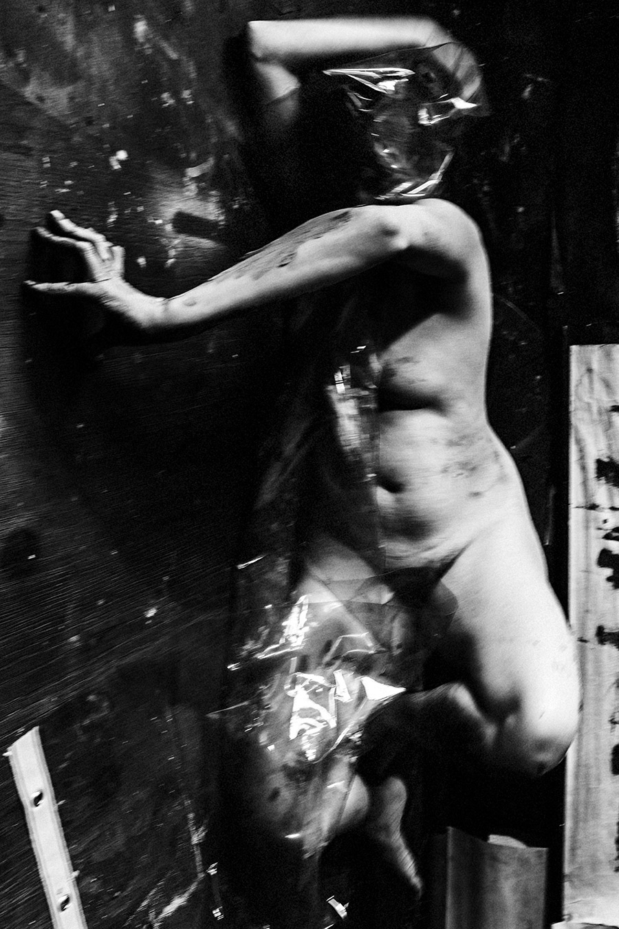 Stehender nackter Körper lehnt an dunkler Wand, Kopf durch Folie verdeckt, eine Hand ausgestreckt, Ton auf der Haut.