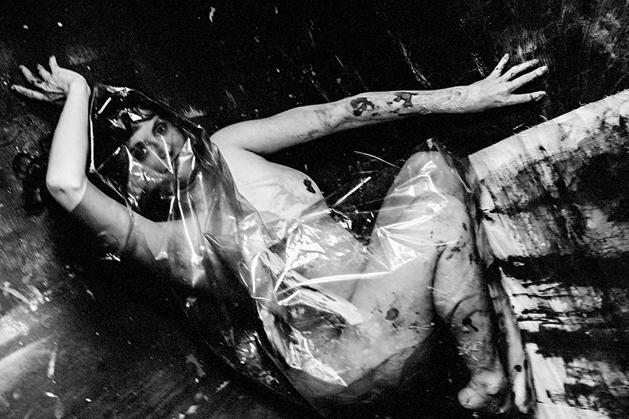 Kniende Frau, Oberkörper von ton-verschmiertem schwarzem Stoff eingehüllt, Gesicht verborgen, Bauch und Beine frei.