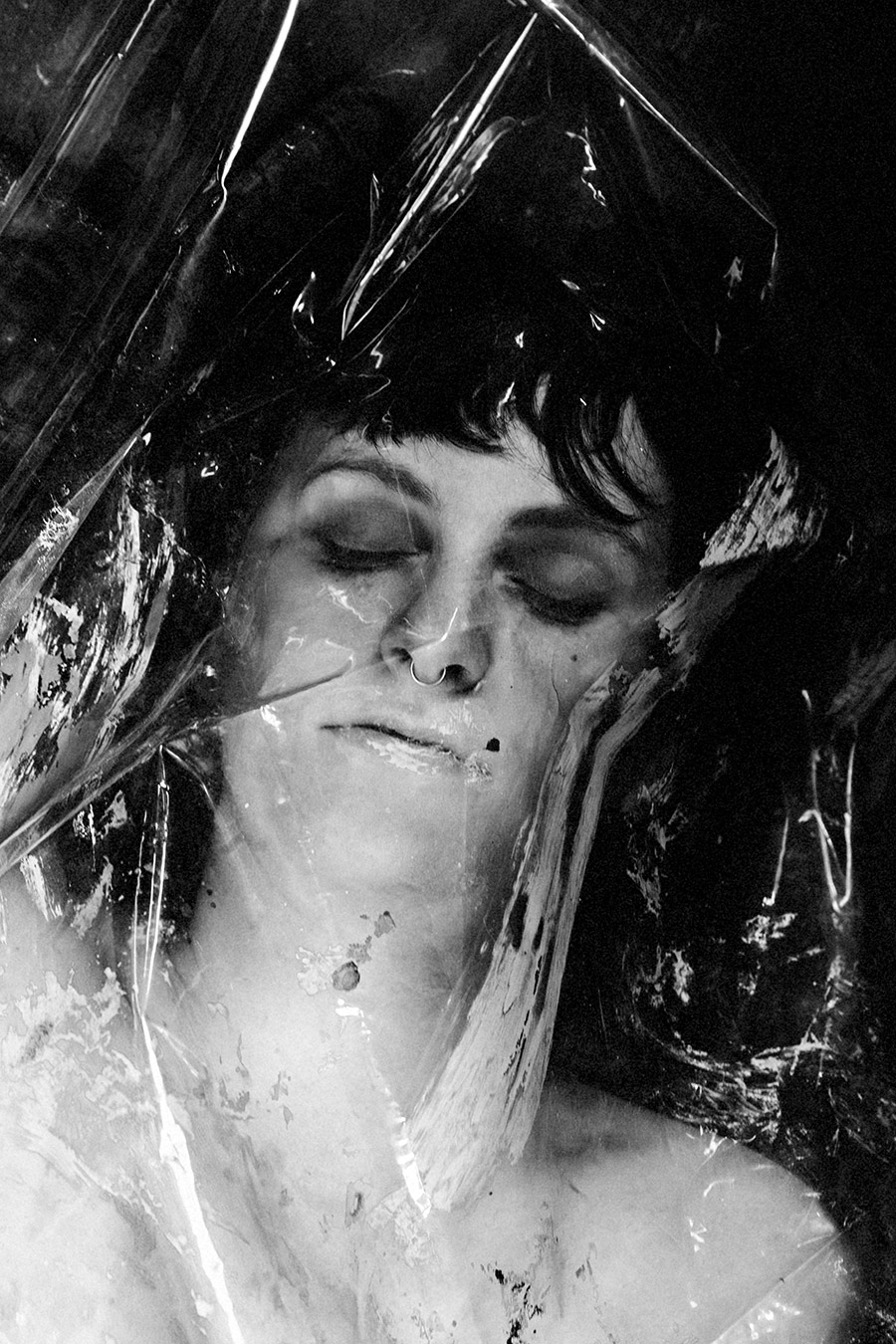 Gesicht einer Frau hinter zerknitterter Klarsichtfolie, Augen geschlossen, Ton- und Fettspuren auf der Haut.
