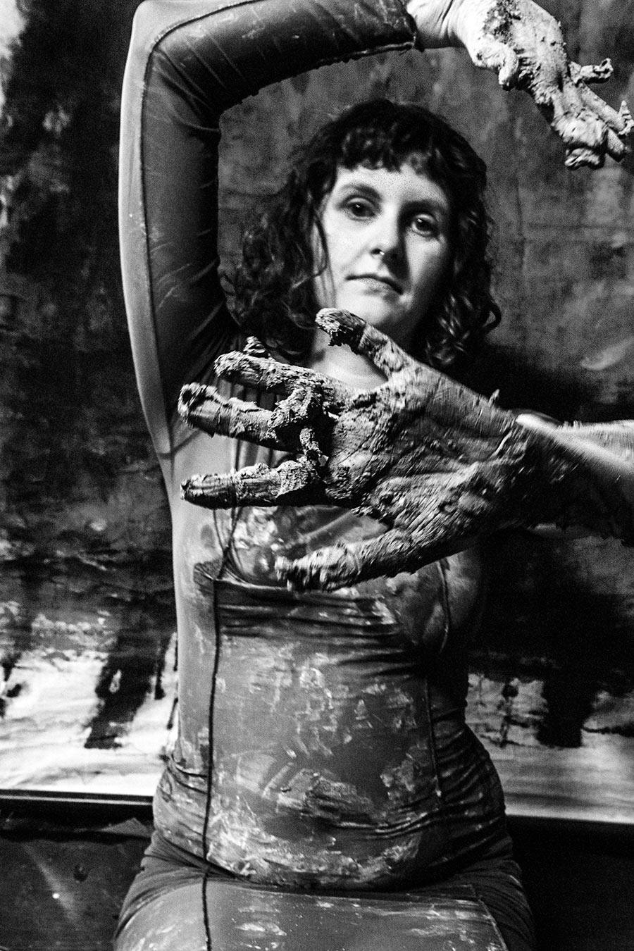 Schwarzweißfoto: Frau im ton-bedeckten Kleid blickt direkt in die Kamera, eine ton-verkrustete Hand vor dem Körper ausgestreckt, Arm über den Kopf gehoben, abstrakter Hintergrund.