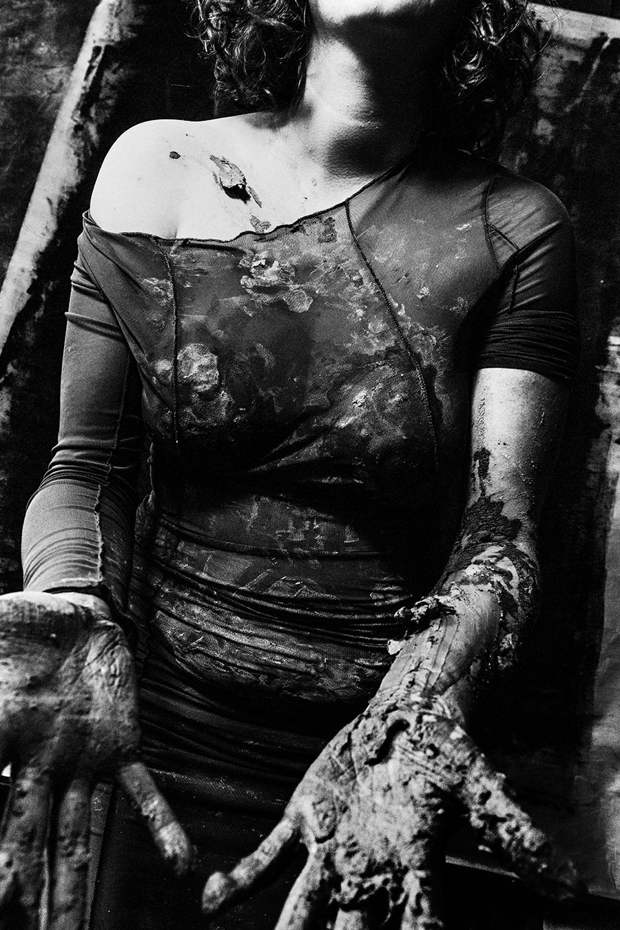 Oberkörper einer Frau im ton-bedeckten transparenten Kleid, Hände und Unterarme dicht mit getrocknetem Ton überzogen, Schulter frei, Kopf angeschnitten.