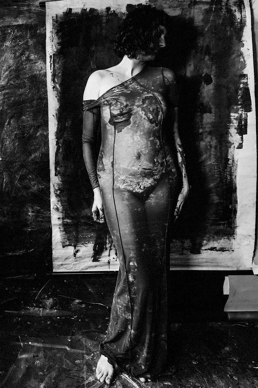 Stehende Frau in engem transparentem Kleid, das mit Ton bedeckt ist, vor großformatigem abstraktem Gemälde. Kopf geneigt, Blick gesenkt.
