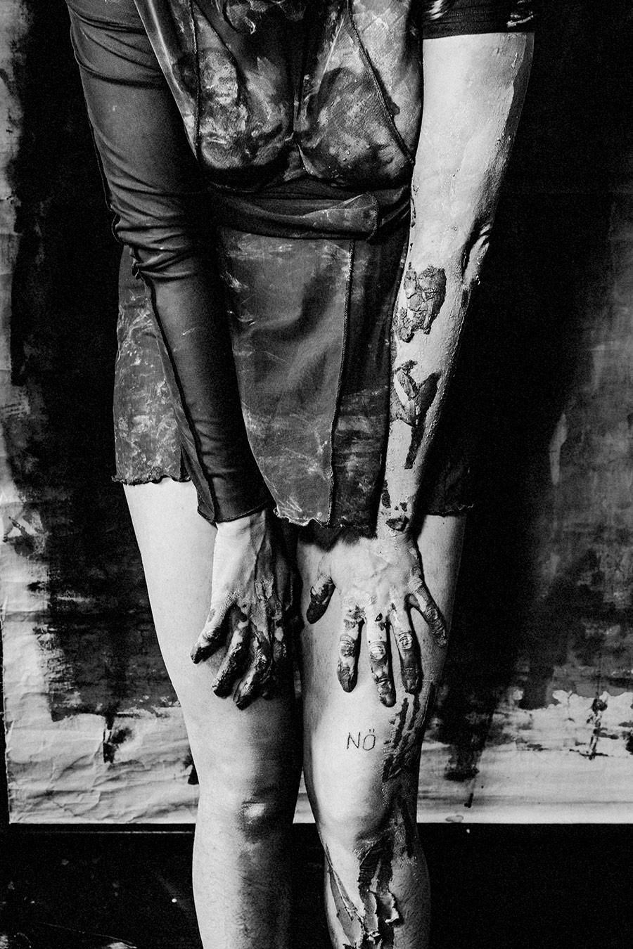 Schwarzweißfoto: Nahaufnahme von Unterschenkeln und ton-bedeckten Händen, die auf den Knien ruhen. „NÖ"-Tattoo auf dem Schienbein sichtbar.