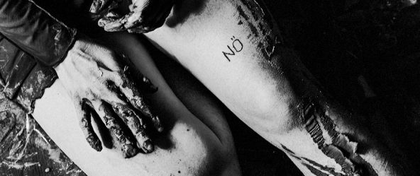 Alttext: Schwarzweißnahaufnahme: Zwei ton-bedeckte Hände auf einem nackten Oberschenkel mit „NÖ"-Tattoo, Holzboden mit Farbresten im Hintergrund.
