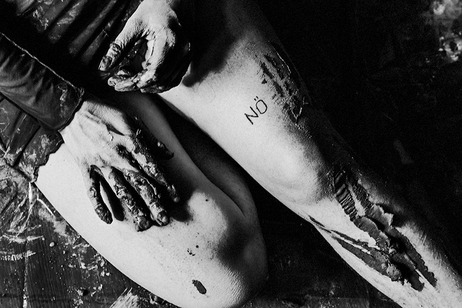 Alttext: Schwarzweißnahaufnahme: Zwei ton-bedeckte Hände auf einem nackten Oberschenkel mit „NÖ"-Tattoo, Holzboden mit Farbresten im Hintergrund.