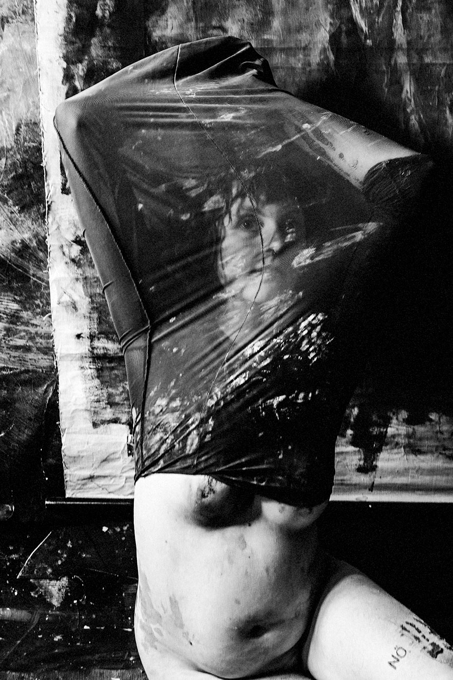 Schwarzweißfoto: Kniende Frau, Kopf tief gesenkt, Gesicht von transparentem Material bedeckt, Ton auf Schultern und Armen.