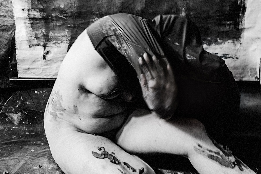 Schwarzweißfoto: Kniende Frau, Oberkörper von ton-verschmiertem schwarzem Stoff eingehüllt, Gesicht verborgen, Bauch und Beine frei.