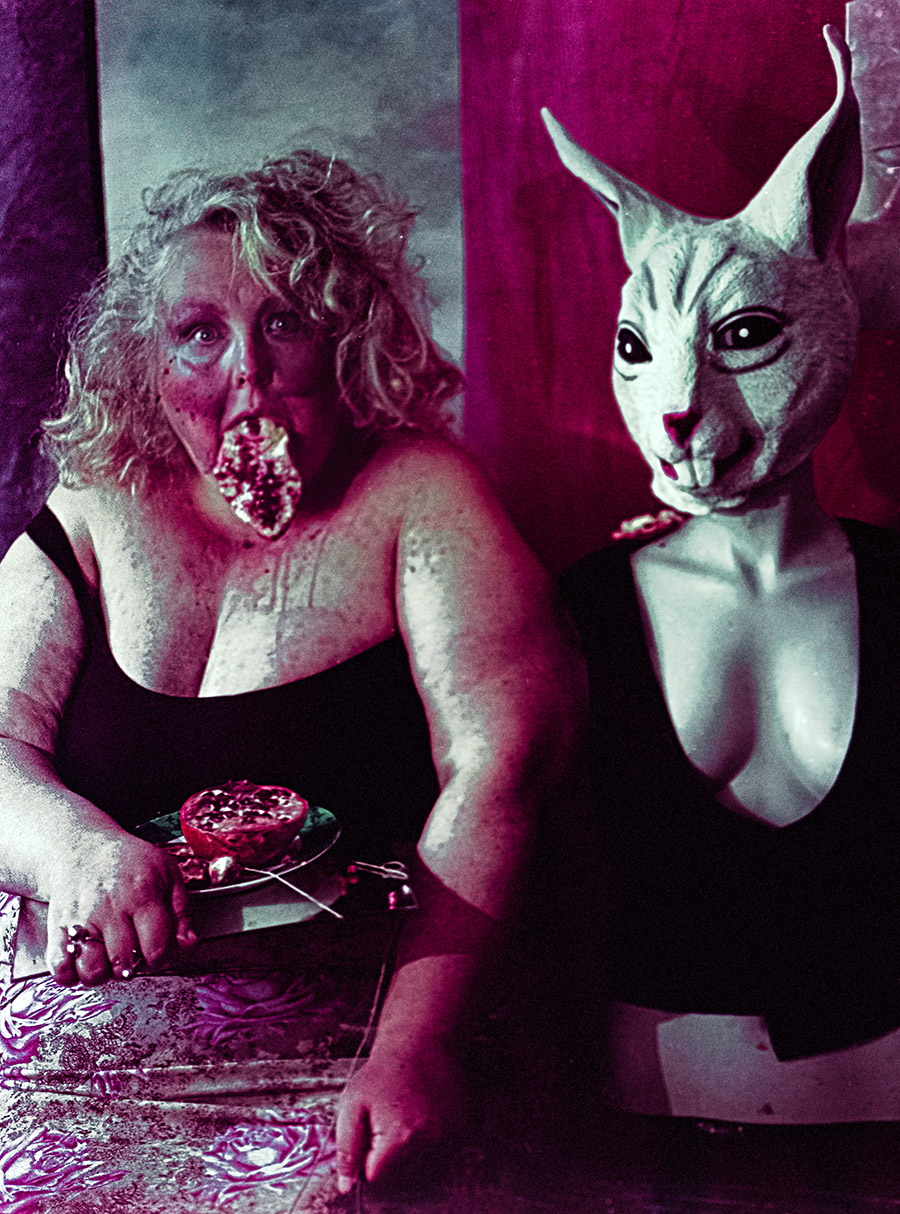 Frontalaufnahme beider Figuren, Frau mit Granatapfel im Mund, Häschenmaske daneben, gedeckter Tisch mit aufgeschnittenem Granatapfel und Besteck, intensive Filmsuppe-Farbverschiebung — analoge Mittelformat-Fotokunst Antje Kröger Leipzig, Serie Tischgesellschaft