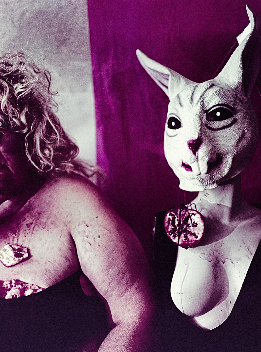 Hochformat, Häschenmaske mit blutendem Granatapfel-Anhänger im Vordergrund, Frau mit Granatapfelstück auf der Schulter im Hintergrund, starke Magenta-Violett-Farbverschiebung durch Filmsuppe — Mittelformat Fotokunst Antje Kröger, Serie Tischgesellschaft