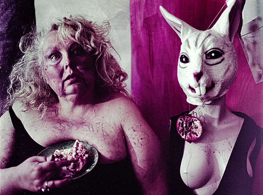 Zwei Figuren am Tisch, eine hält einen Teller mit Granatapfel, die andere trägt Häschenmaske mit Granatapfel-Anhänger, violette Farbverschiebung auf Kodak Color 200 — Antje Kröger Fotokunst Leipzig Berlin, Serie Tischgesellschaft