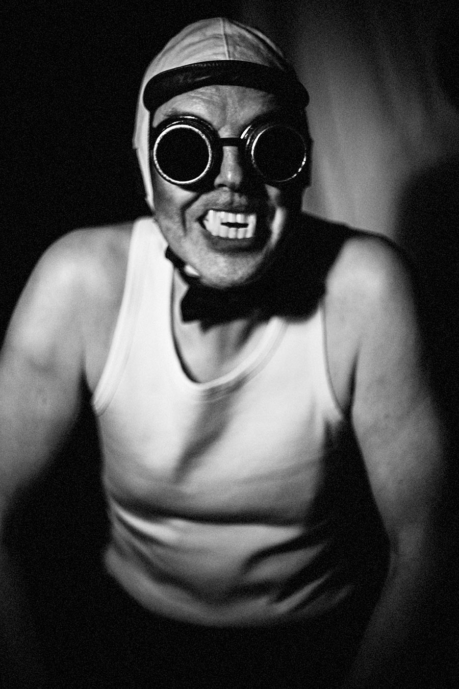 Schwarzweißporträt: Mann mit Schweißerbrille, Lederkappe und Vampirzähnen lacht breit in die Kamera, nach vorne gebeugt, Unterhemd, Fotokunst Antje Kröger