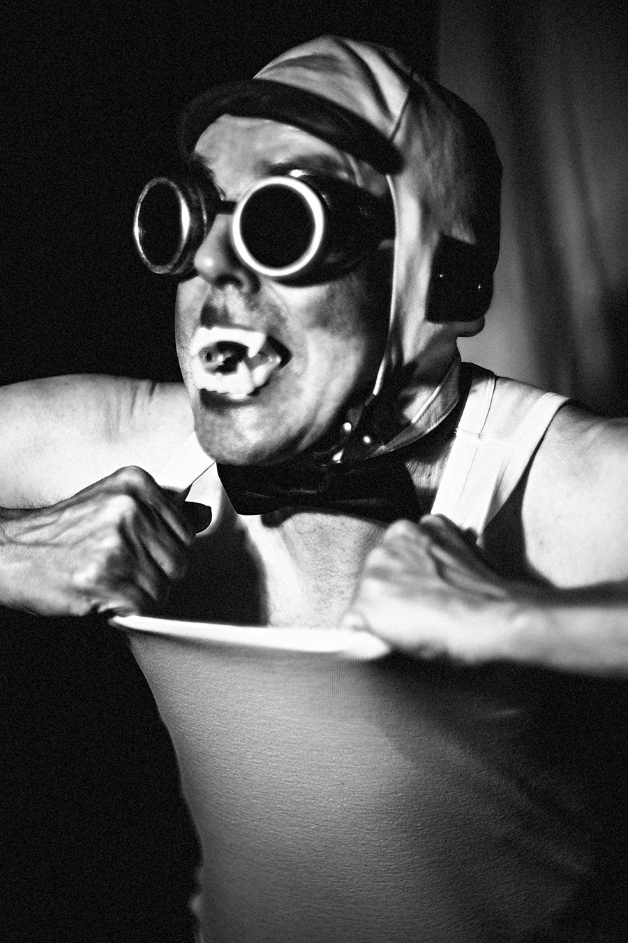 Schwarzweißporträt: Mann mit Schweißerbrille, Lederkappe, Vampirzähnen und Fliege will sein Unterhemd zerreißen, expressiver Ausdruck, Fotokunst Antje Kröger