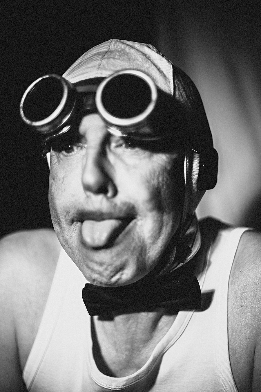 Schwarzweißporträt: Mann mit Schweißerbrille und Lederkappe streckt die Zunge heraus, Fliege, Unterhemd, Nahaufnahme, Fotokunst Antje Kröger