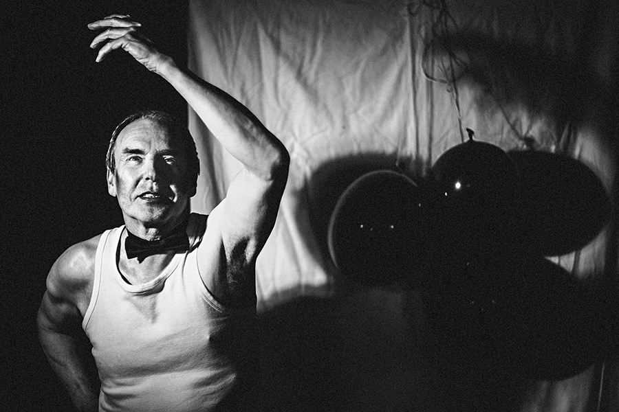 Schwarzweißfotografie: Älterer Mann im Unterhemd mit Fliege, hebt den Arm, schwarze Luftballons im Hintergrund, dramatisches Licht, Fotokunst Antje Kröger