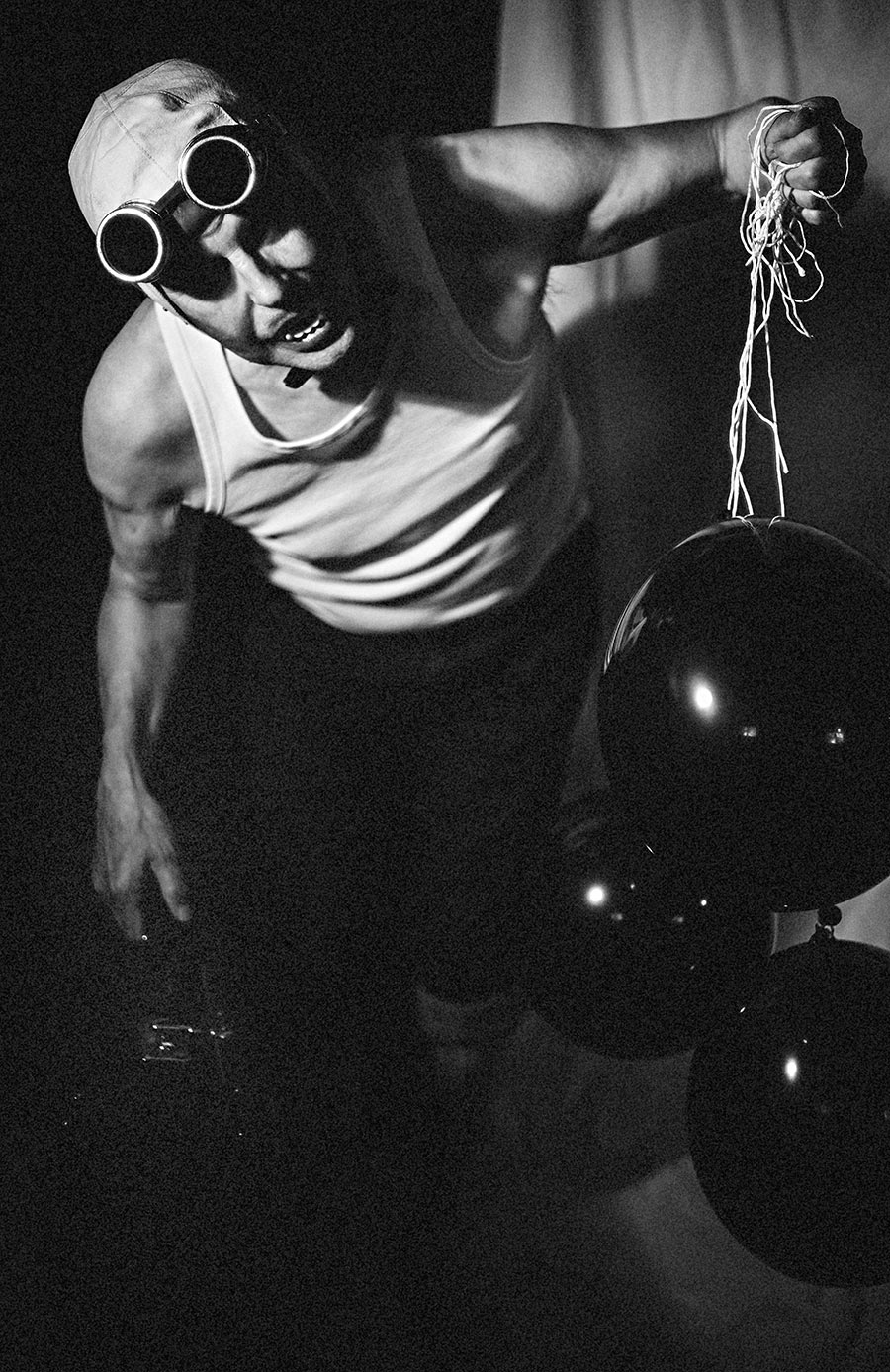 Schwarzweißfotografie: Mann mit Schweißerbrille und Vampirzähnen hält ein Bündel Schnüre schwarzer Luftballons, dynamische Körperhaltung, Unterhemd, Fotokunst Antje Kröger