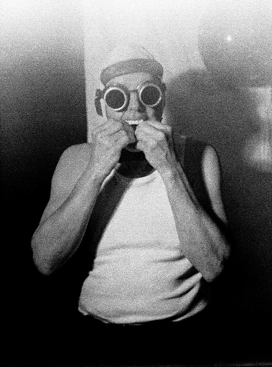 Analoges Schwarzweißporträt: Mann mit Schweißerbrille und Vampirzähnen hält sich beide Hände vors Gesicht, tiefes Schwarz, Mamiya Mittelformat, Antje Kröger