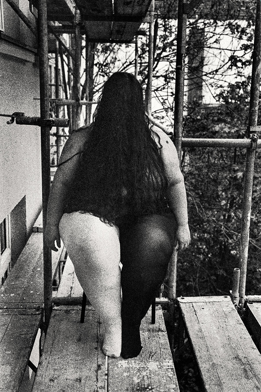 Sitzende Figur auf Gerüst, Frühling, langes Haar, dicker Körper gegen kahle Bäume, Schwarz-weiß Analogfoto – Fotokunst Antje Kröger Leipzig Berlin, Serie Walsturz