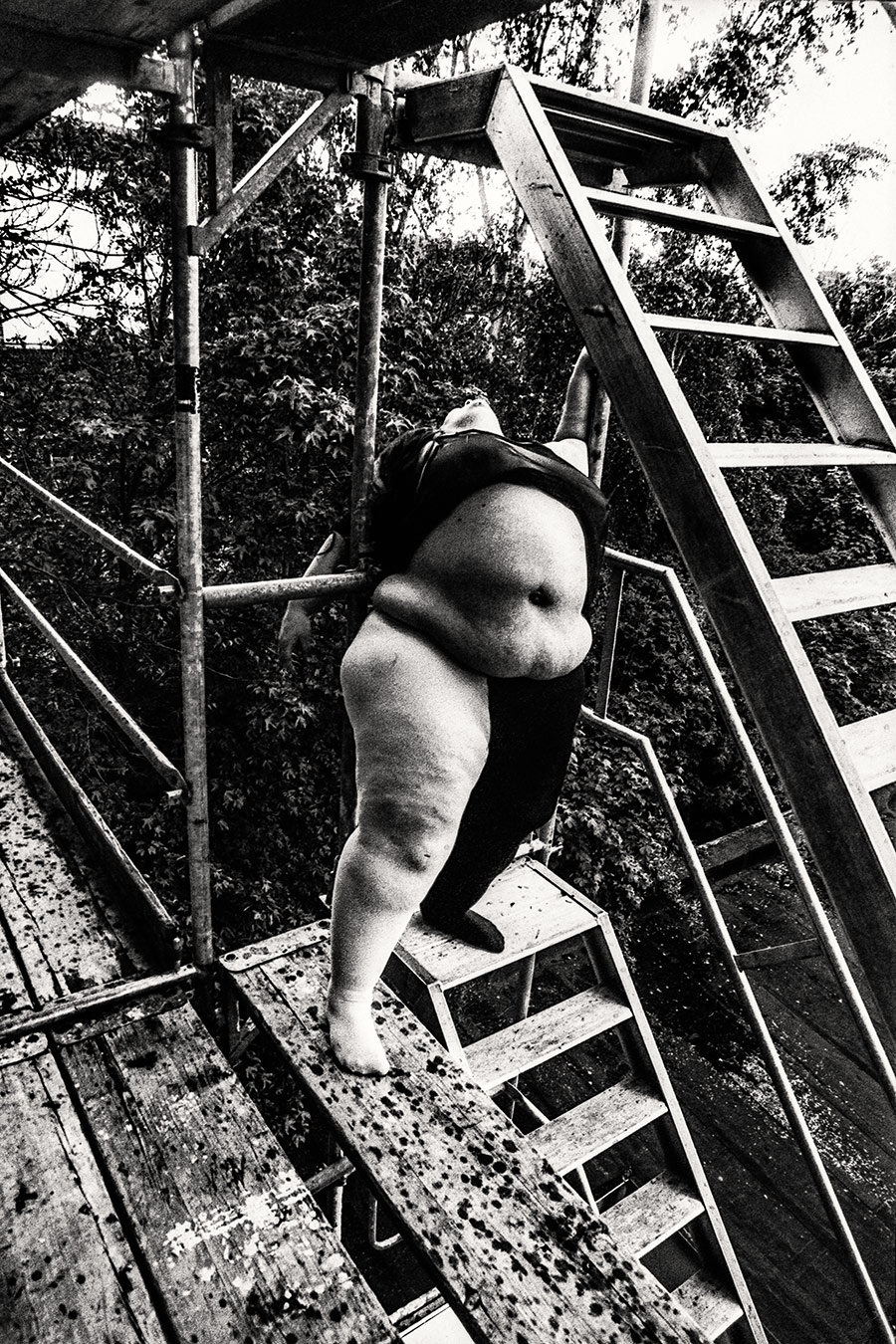 Dicke Frau hängt rückwärts an Gerüstleiter, Kopf zurückgeworfen, Stadtdächer im Hintergrund, extremes 35mm-Korn, analoges Schwarzweiß – Antje Kröger Fotokunst Berlin Leipzig, Serie Walsturz