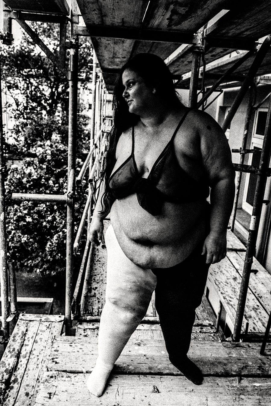 Dicke Frau stehend auf Gerüst im Sommer, Blick zur Seite, Binde am Bein, analoges 35mm-Schwarzweiß – Antje Kröger Fotokunst Leipzig Berlin, Serie Walsturz