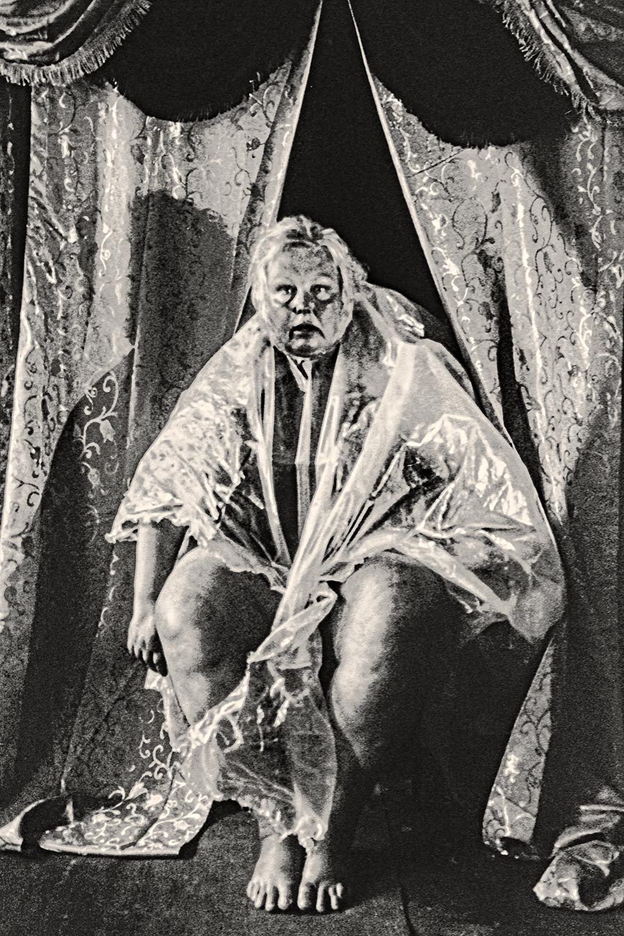 Analoge Fotografie einer sitzenden Frau in transparenter Folie zwischen geöffneten Theatervorhängen, Fomapan Ortho 400 gepusht