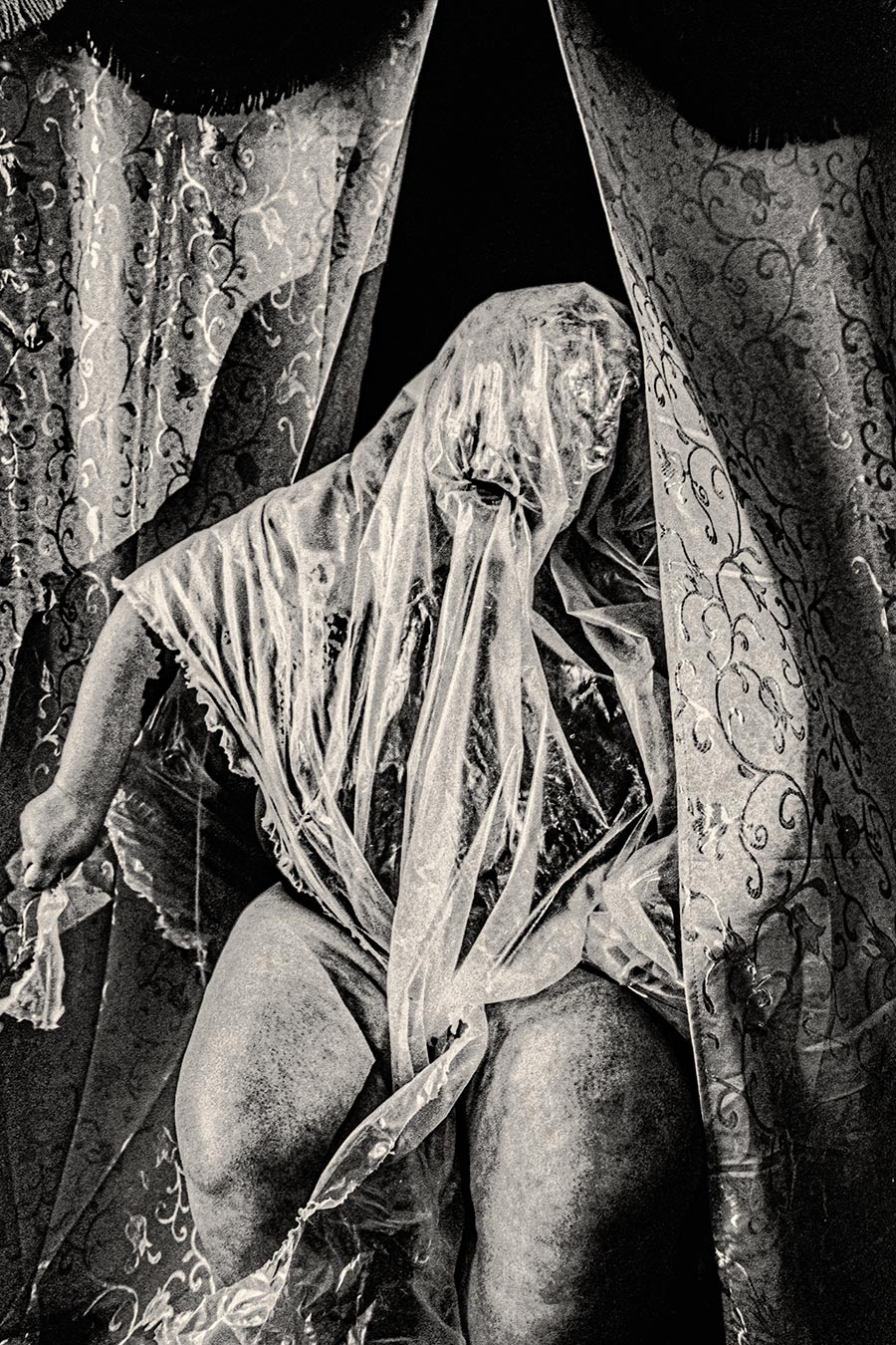 Person mit transparenter Folie über dem Kopf zwischen gemusterten Vorhängen, analoges Schwarzweißfoto Fomapan Ortho 400 gepusht