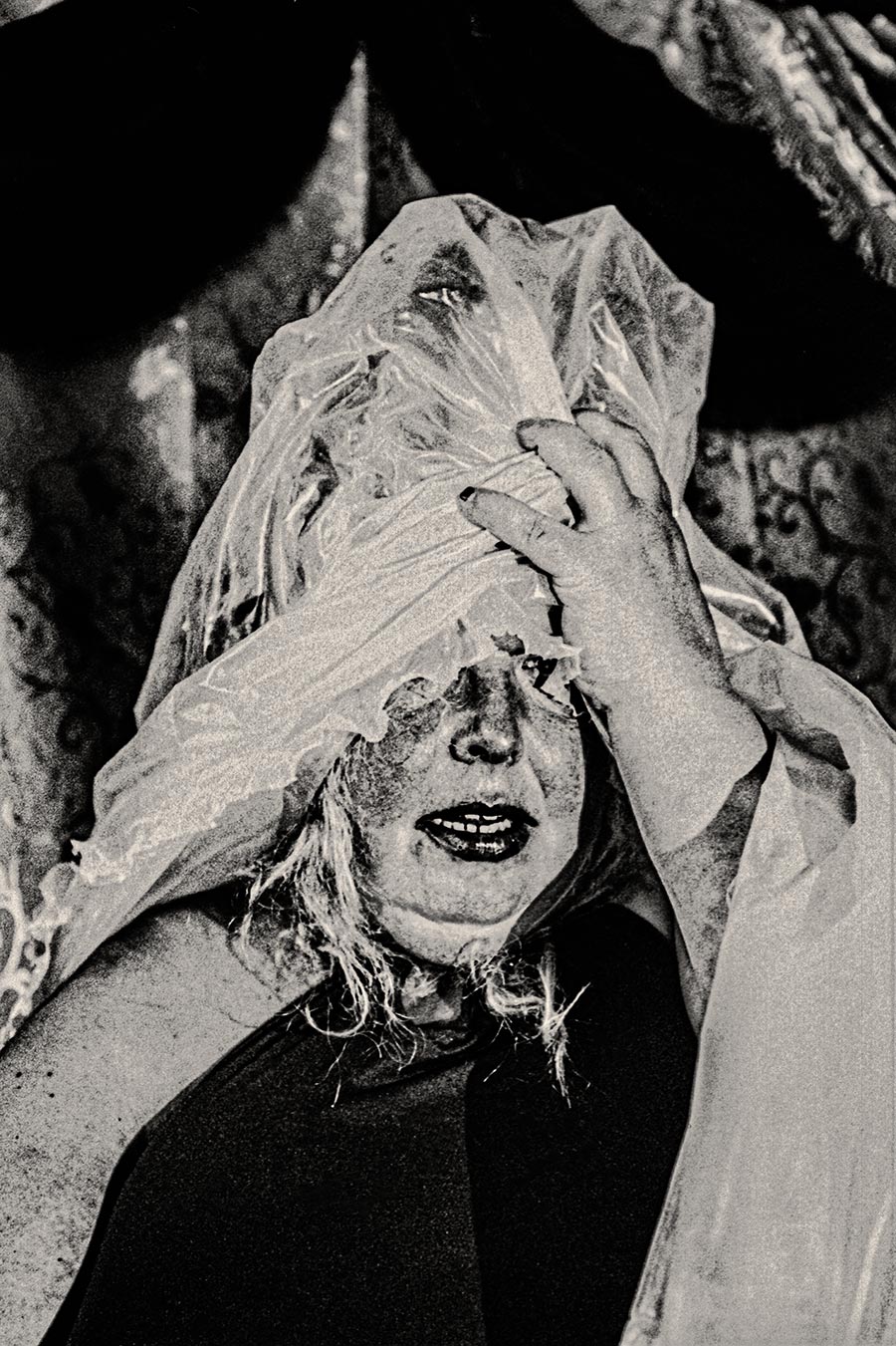 Frau hält transparente Folie vor ihr Gesicht, Mund geöffnet, dramatisches analoges Schwarzweißfoto mit starkem Korn