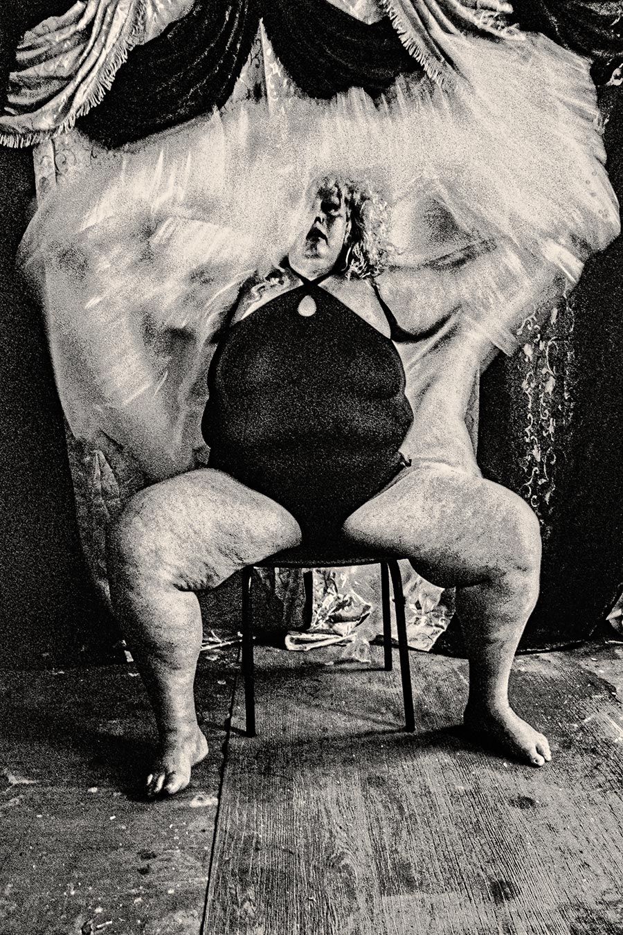 Dickere Frau sitzt auf Hocker im schwarzen Badeanzug, weißer Tüll bewegt sich über ihr, analoges Schwarzweißfoto Fomapan Ortho 400