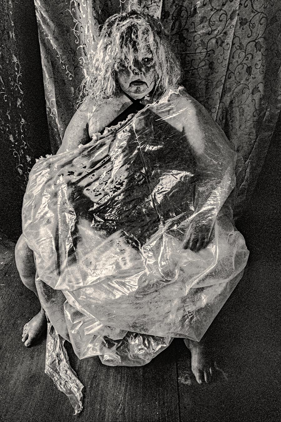 Frau auf dem Boden sitzend, von transparenter Folie umgeben, Gesicht mit schwarzer Schminke, direkter Blick in die Kamera, analoges Schwarzweißfoto