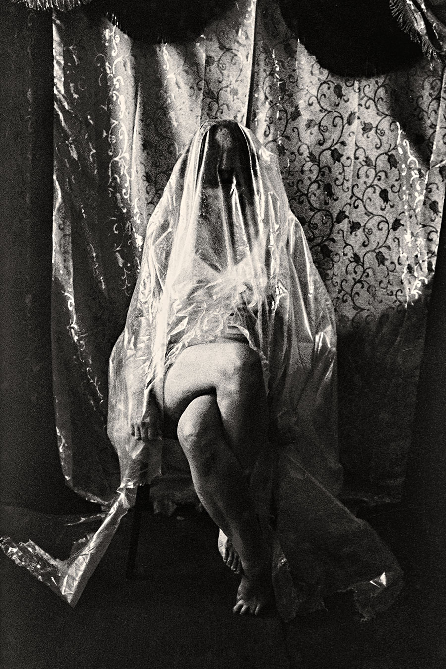Sitzende Person mit transparenter Folie als Umhang, ein Bein frei, Gesicht nicht sichtbar, analoges Schwarzweißfoto Fomapan Ortho 400 gepusht