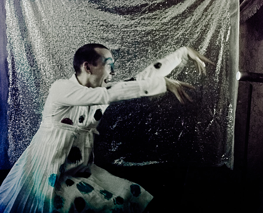 Ausdrucksstarke Bewegung vor Pailletten-Vorhang, Mann mit ausgestreckten Armen, Mund geöffnet, weißes Kleid mit dunklen Flecken, analoge Performance-Fotografie Meeresschnee 3