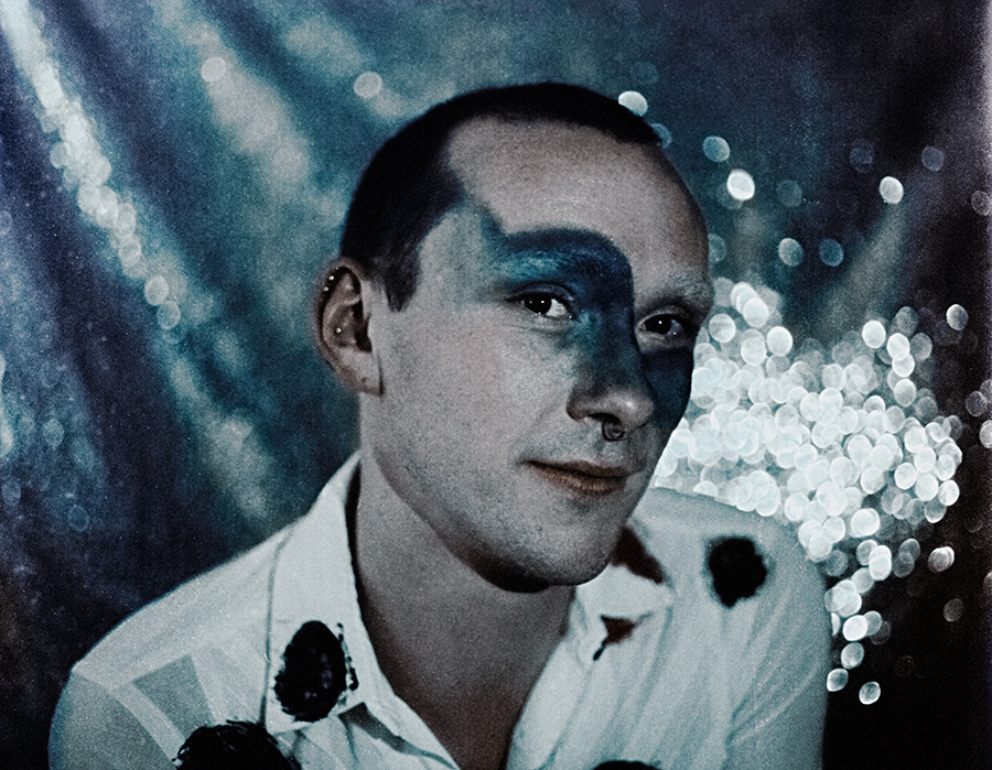 Analoges Porträt Meeresschnee 3, Mann mit blauem Augen-Make-up, Pailletten-Hintergrund silber-blau, weißes Hemd mit schwarzen Farbflecken, Fuji Provia 400, Mamiya645, analoge Farbfotografie Antje Kröger
