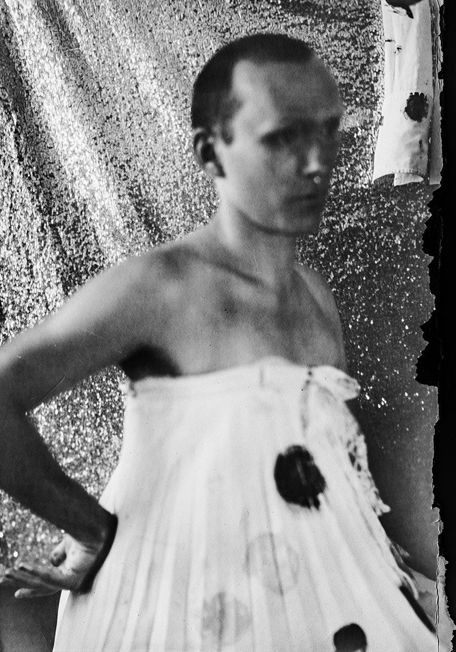 Schwarzweiß-Porträt Kamera Mentor, Mann mit trägerlosem geplisseertem Kleid, schwarze Farbflecken auf Stoff, Pailletten-Hintergrund, unscharfe Bildführung, ORWO Fotopapier, analoge Fotografie Meeresschnee 3