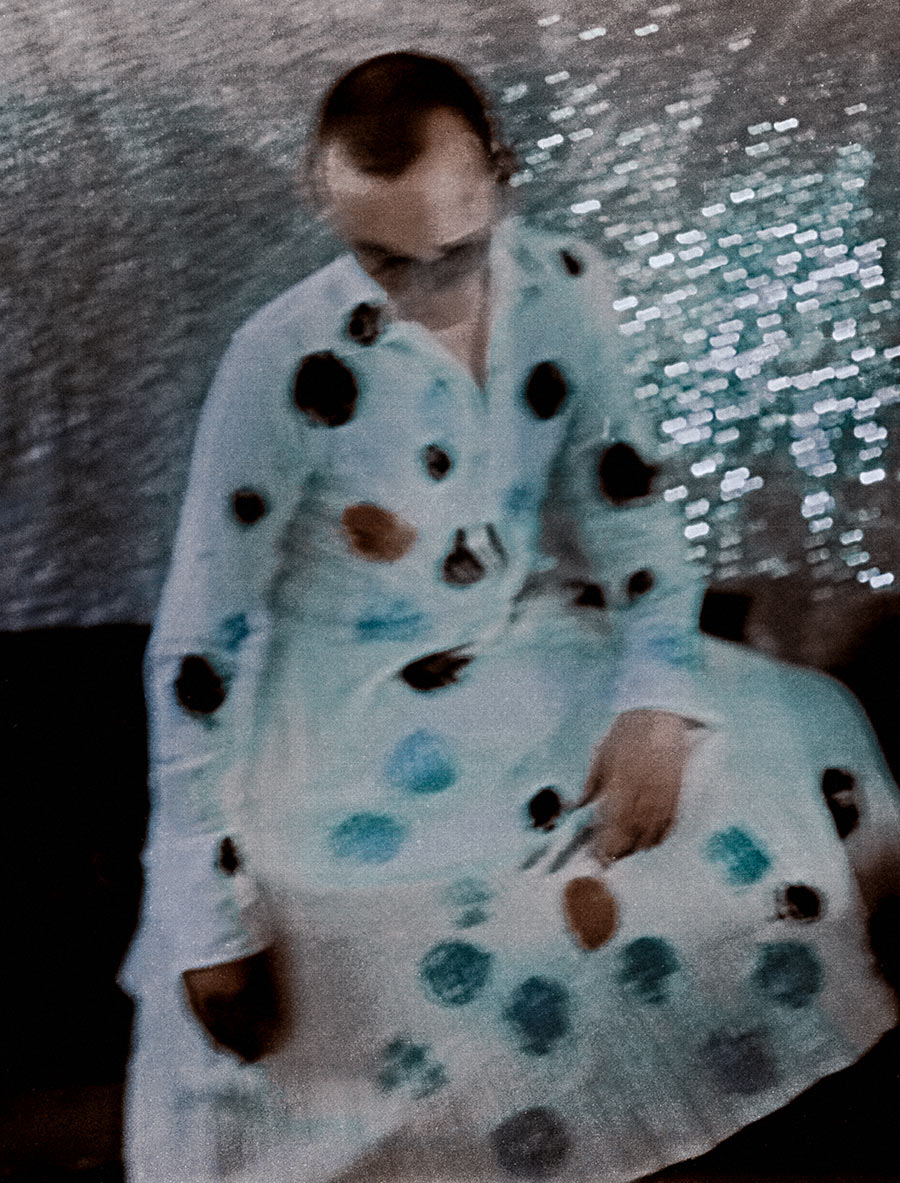Sitzende Person mit gesenktem Blick, weißes Gewand mit bunten Punkten in Rost, Türkis, Schwarz, Glitzerhintergrund grau-silber, analoge Fotografie Meeresschnee 3