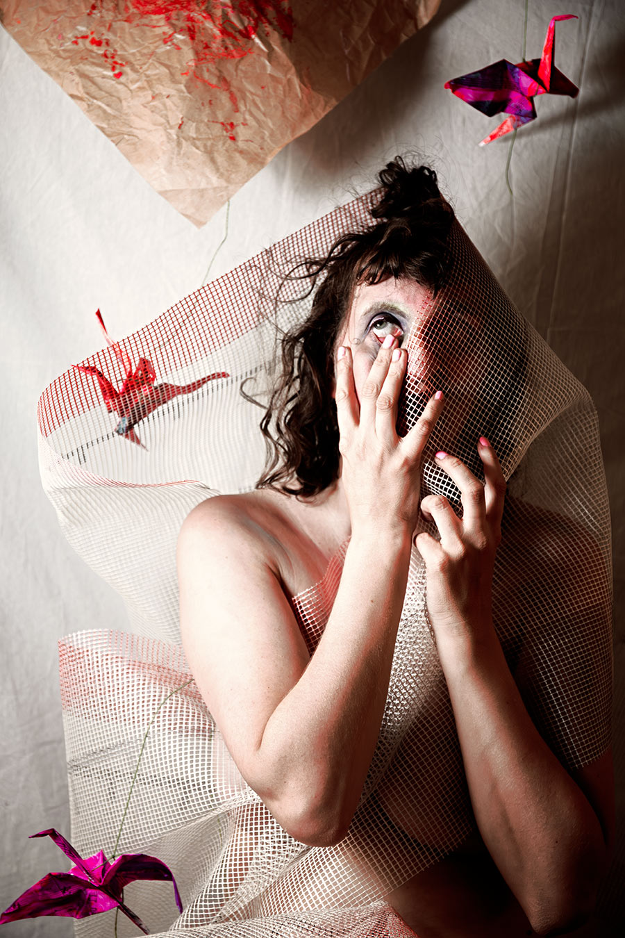 Verdeckte Gestalt mit Hand vorm Gesicht unter Netzstoff, zwei Origami-Kraniche in Rot und Pink, Aufsicht-Perspektive, Canon 6D, inszenierte Fotokunst Vogelfrei