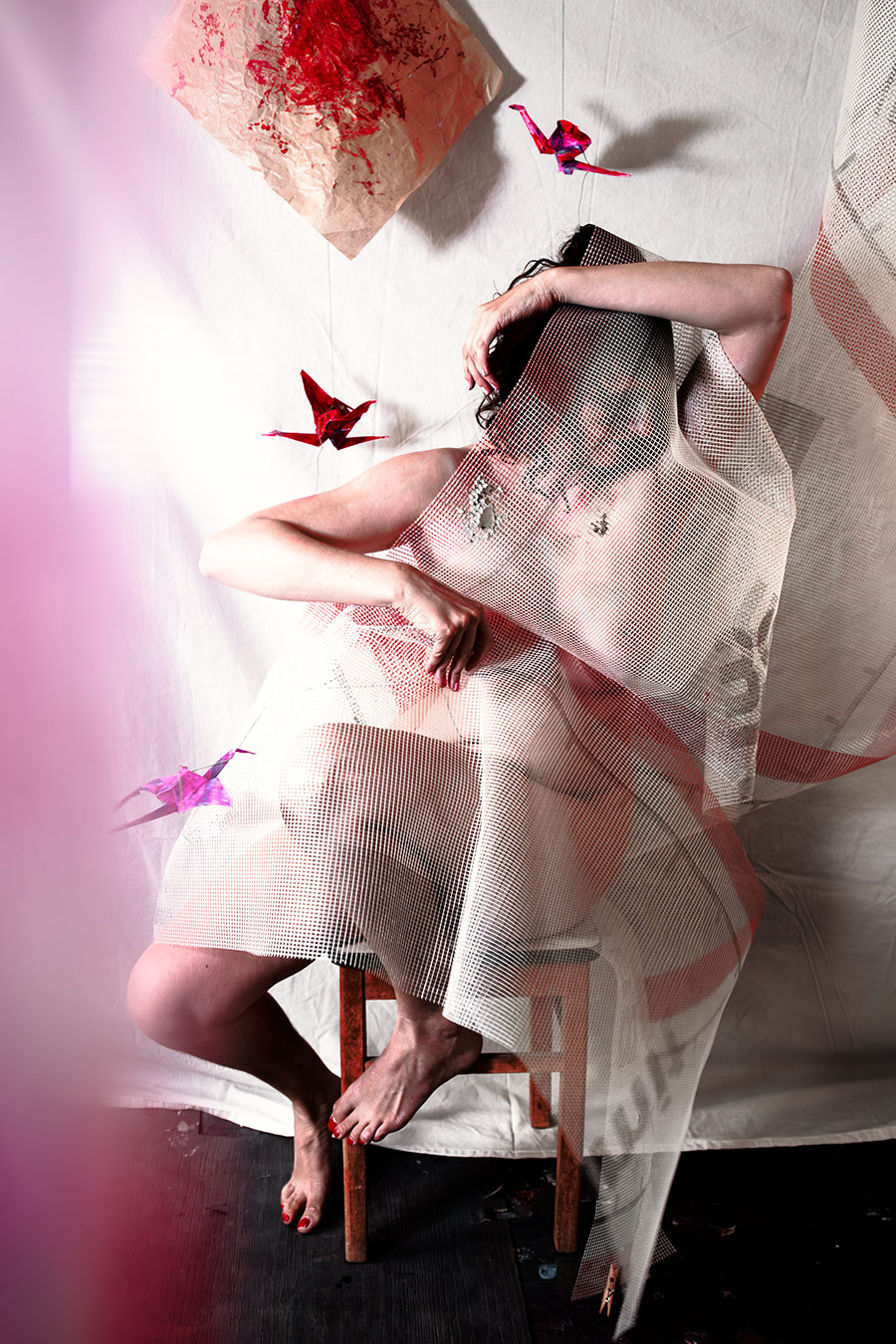 Sitzende Person auf Holzhocker, Netzstoff als Gewand, Origami-Kraniche in Rot und Pink hängend, barfuß mit rotem Nagellack, Canon 6D, konzeptuelle Aktfotografie Vogelfrei