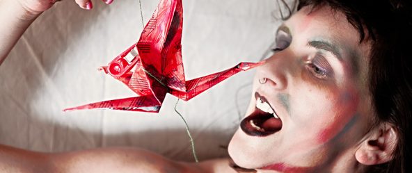 Nahaufnahme Gesicht mit geöffnetem Mund und rotem Origami-Kranich an Faden gehalten, Bodypaint-Schminke, Canon 6D, symbolische Fotokunst Vogelfrei
