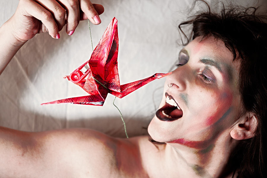 Nahaufnahme Gesicht mit geöffnetem Mund und rotem Origami-Kranich an Faden gehalten, Bodypaint-Schminke, Canon 6D, symbolische Fotokunst Vogelfrei