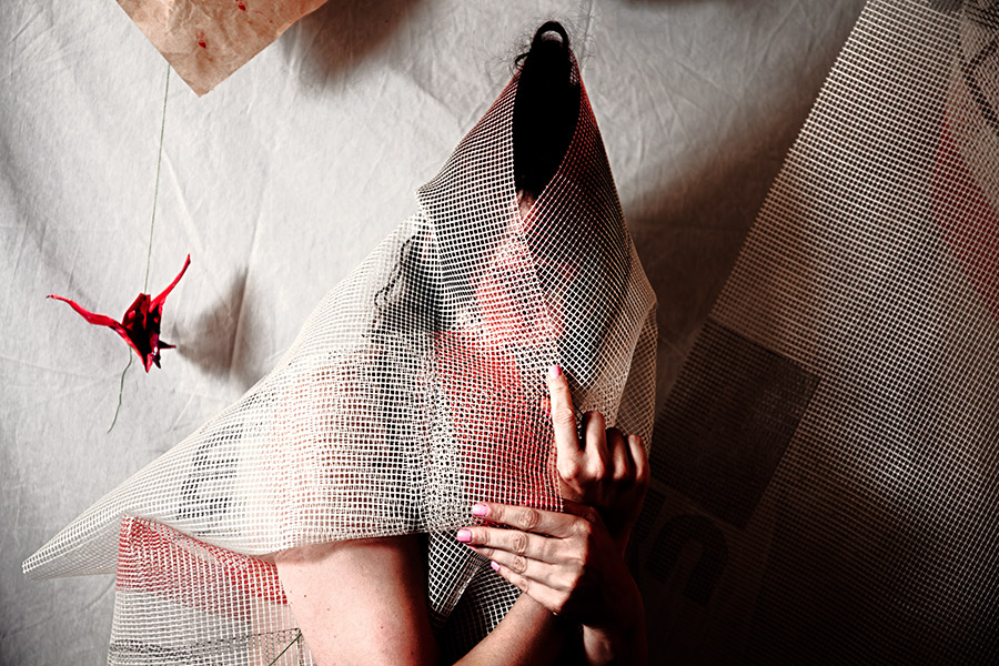 Verhüllte Gestalt unter rot-weißem Netzstoff, Hand hält Stoff fest, Origami-Kranich im Hintergrund, Aufsicht-Perspektive, Canon 6D, Fotokunst Vogelfrei