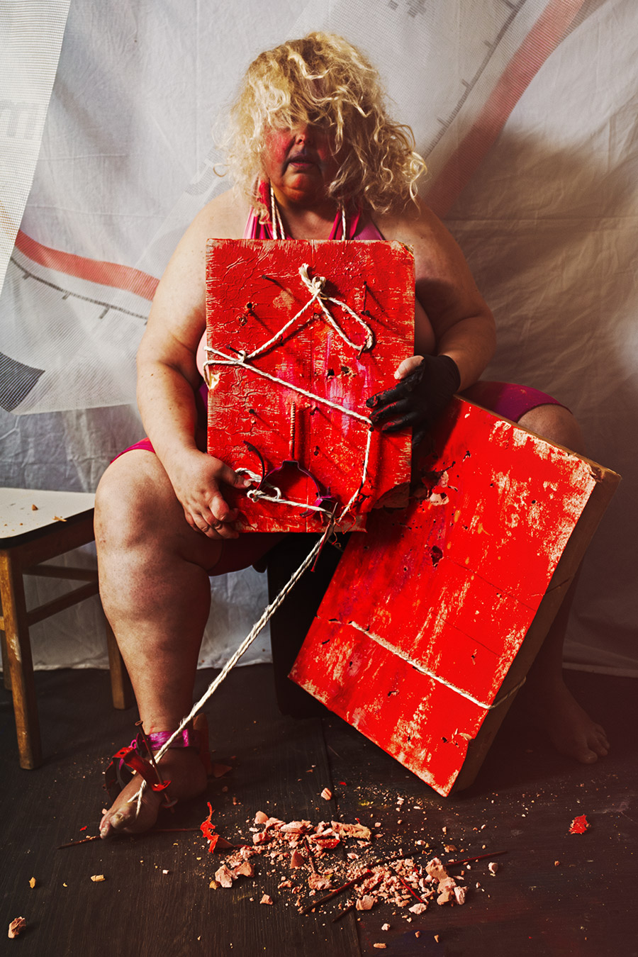 Sitzende Person mit blondem Lockenhaar hinter rotem Holzbrett, schwarzer Handschuh, gefesselter Fuß, Canon 6D, konzeptuelle Bondage-Fotografie Vogelfrei
