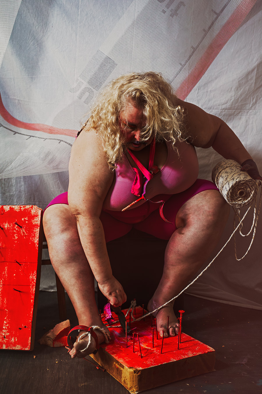 Gebeugte Person bindet Schnur um gefesselten Fuß auf rotem Nagelbrett, Garnrolle in der Hand, Canon 6D, prozesshafte Bondage-Fotografie Vogelfrei