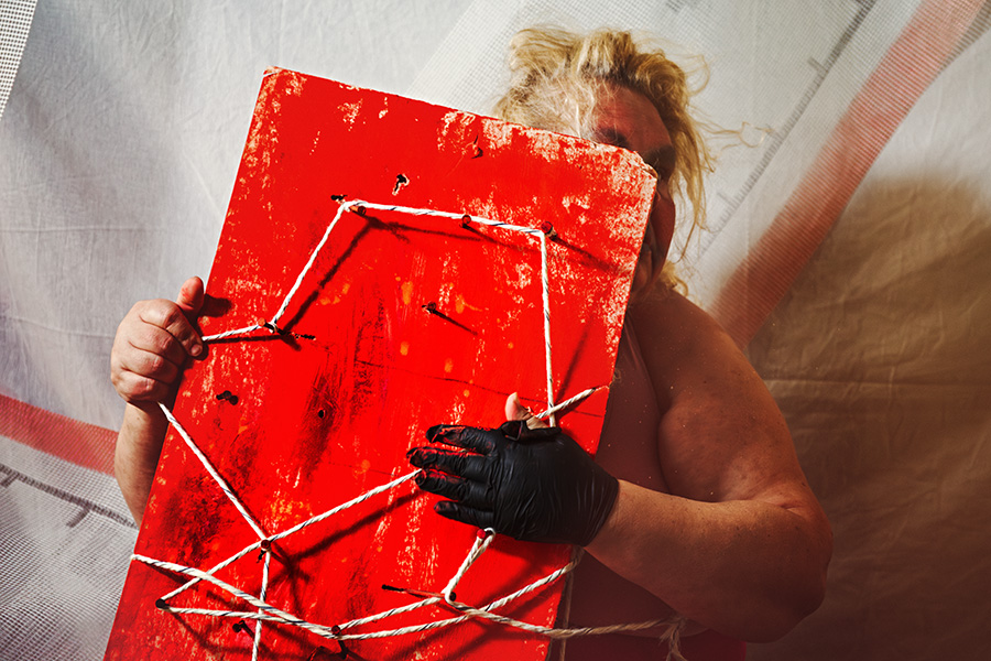 Person hält rotes Nagelbrett mit Schnurmuster vor Körper, schwarzer Handschuh, blondes Haar verdeckt Gesicht, Canon 6D, Bondage-Fotografie Vogelfrei