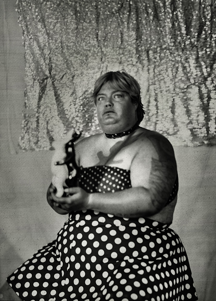 Frau mit dickem Körper hält Porzellanfigur zweier sich festhaltender Häschen in Weiß und Gold, schwarzes Halsband, gepunkteter Rock, glitzernder Hintergrund, clowneske Fine-Art-Fotografie