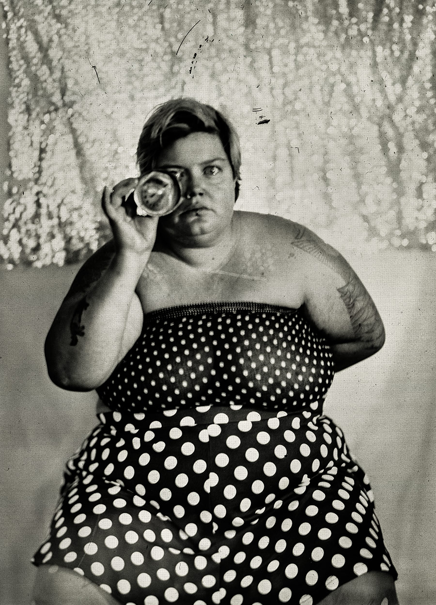 Frau hält ein Glas vor ihr Auge wie ein improvisiertes Fernrohr, gepunktetes Tuch und Rock, glitzernder Hintergrund, analoges Porträt auf Fotopapier, konzeptuelle Fotoserie über Körper und Selbstbild