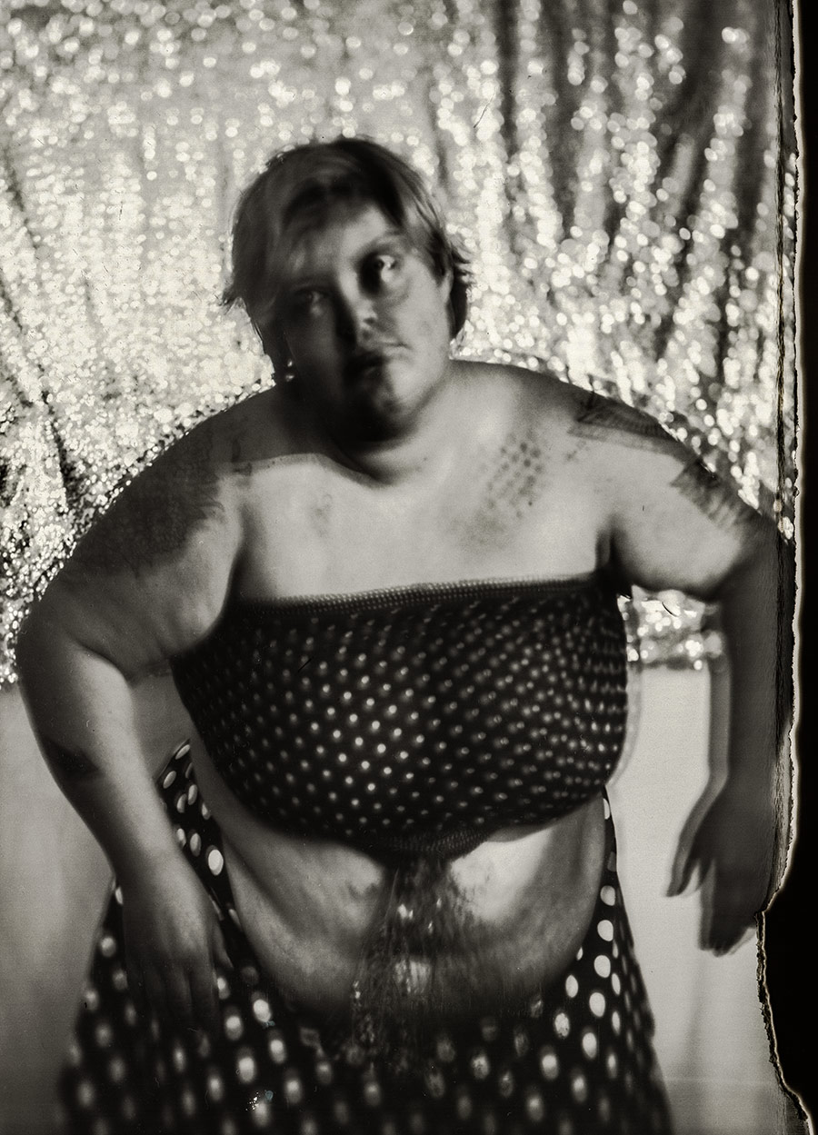 Tätowierte Frau mit dickem, ungeschöntem Körper, gepunktetes Tuch um den Oberkörper geschlungen, glitzernder goldener Hintergrund, analoge Fotografie mit Großformatkamera Mentor, Fine-Art-Porträt