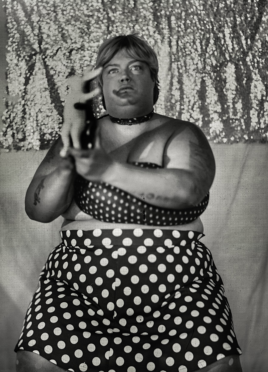 Frau hält Porzellanfigur zweier umschlungener Häschen mit ausgestrecktem Arm vor die Kamera, gepunkteter Rock, Halsband, glitzernder Hintergrund, analoge Großformatfotografie, clowneske Symbolik