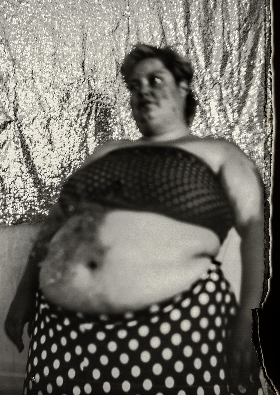 Schwarz-Weiß-Porträt einer tätowierten Frau mit um den Oberkörper geschlungenem gepunktetem Tuch vor glitzerndem Hintergrund, dicker Körper unretuschiert dargestellt, analoge Großformatfotografie mit Kamera Mentor auf Fotopapier, konzeptuelle Body-Positivity-Fotografie