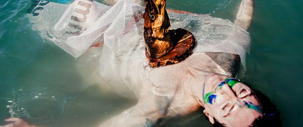 schwimmende figur mit fischkopf-maske treibt rücklings im see, netzstoff um den körper, surreale wasserfotografie fischtanz, pentacon-objektiv m42