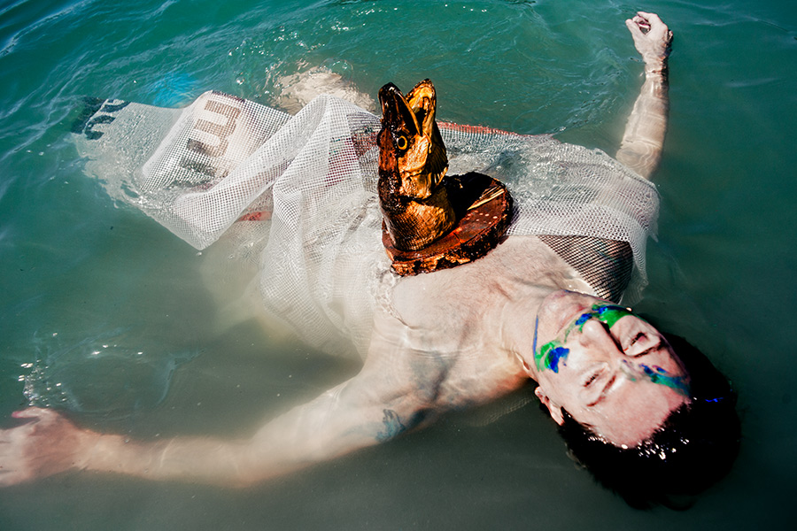 schwimmende figur mit fischkopf-maske treibt rücklings im see, netzstoff um den körper, surreale wasserfotografie fischtanz, pentacon-objektiv m42