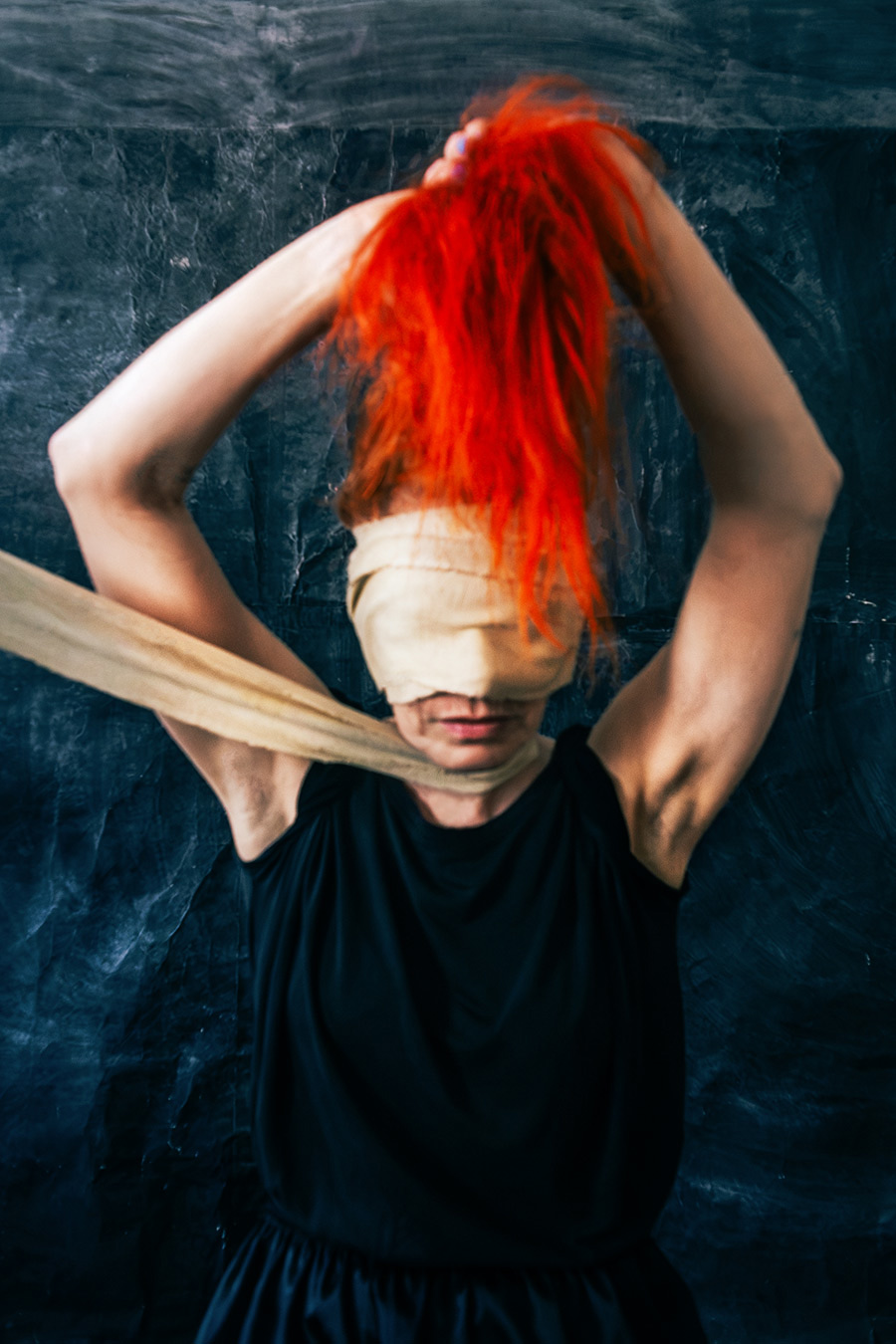 Frau bindet sich selbst eine Augenbinde ab, Arme erhoben, rotes Haar nach oben gerafft, schwarzes Kleid, dunkler Hintergrund, inszenierte Selbstporträt-Fotografie, Thema Imagination und Selbstbestimmung